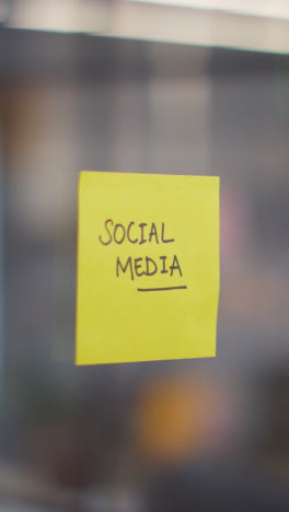 Vertical-Video-Close-Up-Of-Woman-Putting-Sticky-Note-With-Social-Media-Written-On-It-Onto-Transparent-Screen-In-Office